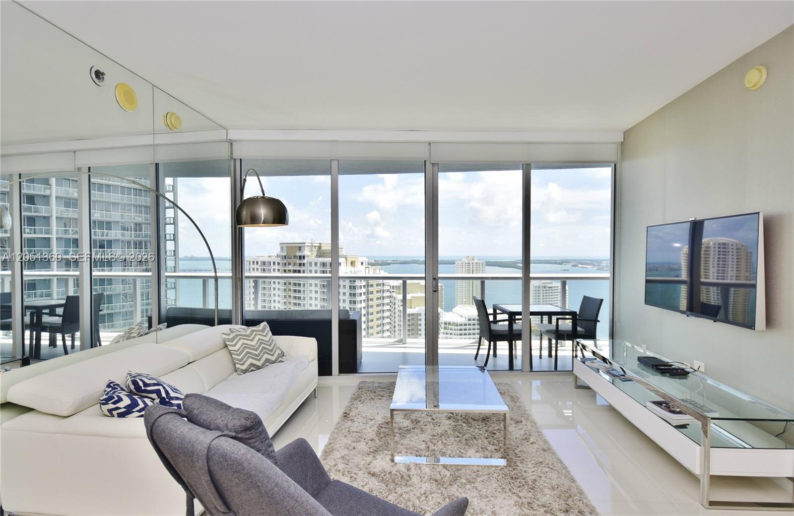 A modern living room features a white sofa, glass coffee table, and floor-to-ceiling windows overlooking water — 465 BRICKELL AVENUE # 3205, Miami FL 33131, MIAMI, FL