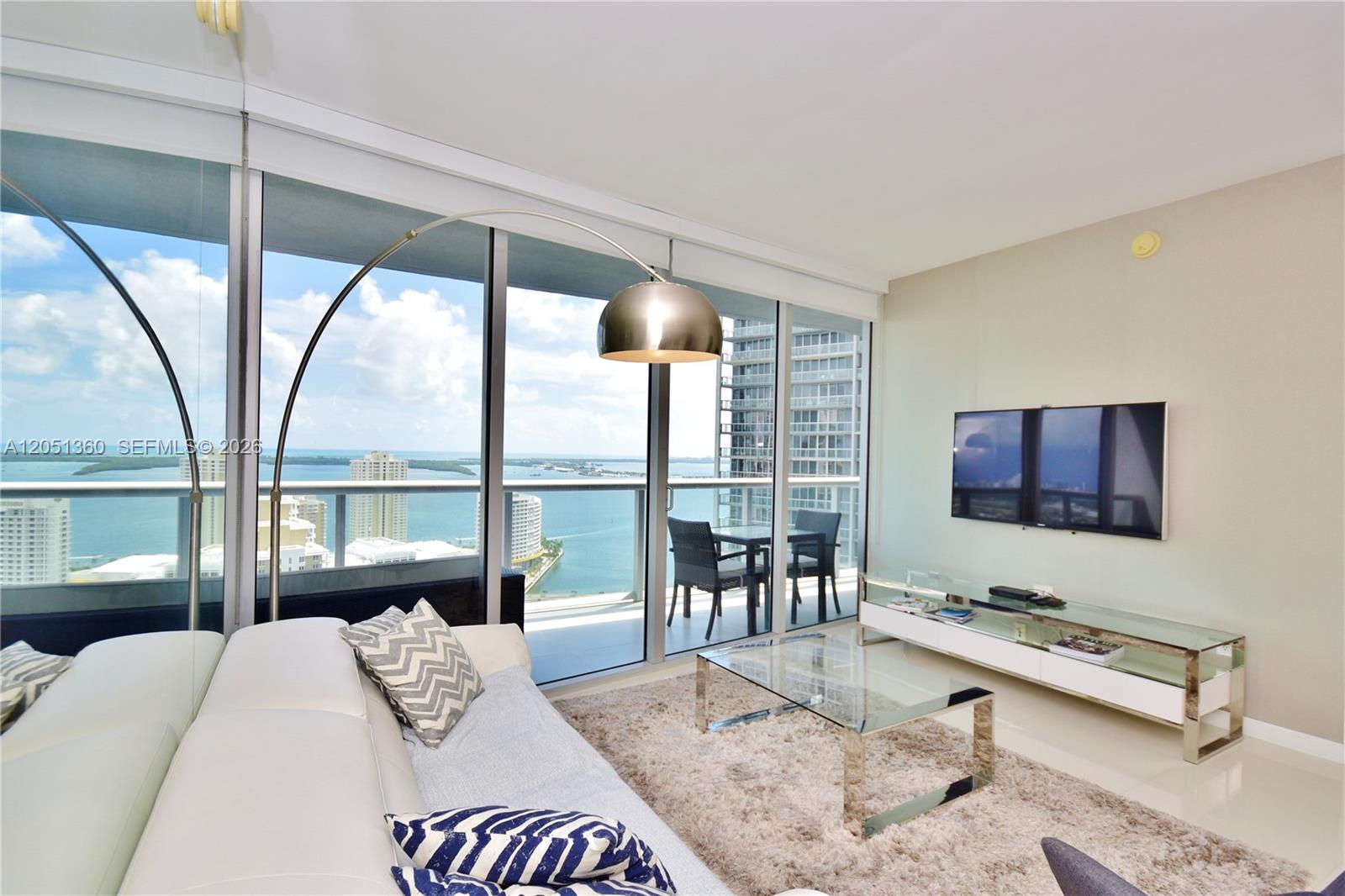 A modern living room features a white sofa, glass furniture, and floor-to-ceiling windows overlooking water — 465 BRICKELL AVENUE # 3205, Miami FL 33131, MIAMI, FL