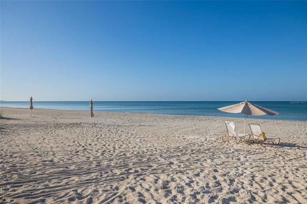 A sandy beach features two closed umbrellas and a set of chairs under an open umbrella — 1095 GULF OF MEXICO DRIVE UNIT 102, LONGBOAT KEY, FL