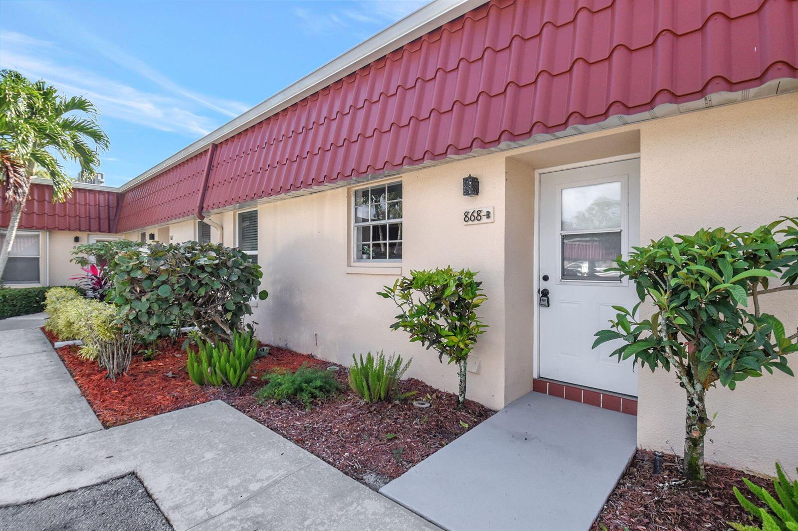 A single-story residential unit features a red metal roof, beige stucco walls, and a white door — 868 Worcester Lane # B, Lake Worth FL 33467, LAKE WORTH, FL