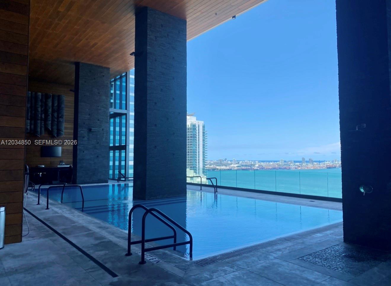 An indoor-outdoor swimming pool with a glass railing overlooking a city skyline and ocean — MIAMI, FL