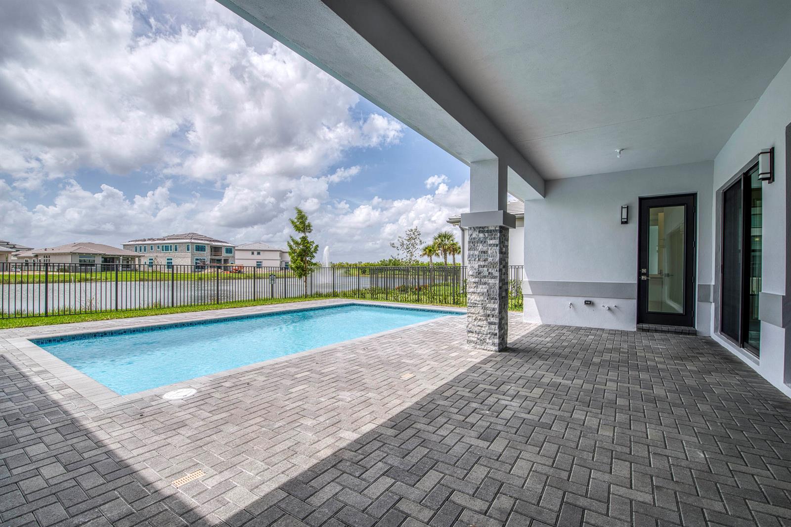 A covered patio with brick pavers overlooks a swimming pool and a fenced lake view — 13559 Nesting Pines Way, Palm Beach Gardens FL 33412, PALM BEACH GARDENS, FL