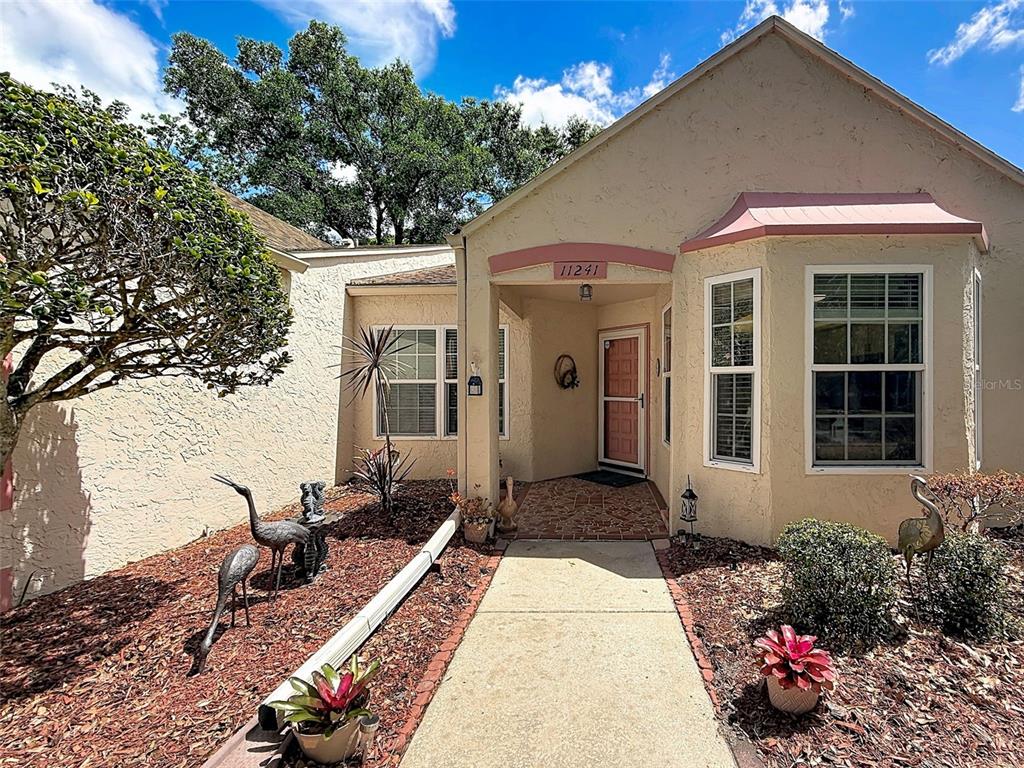 A tan stucco house features a covered entryway, a brick walkway, and landscaped garden beds — 11241 VERSAILLES LANE UNIT 0, PORT RICHEY, FL