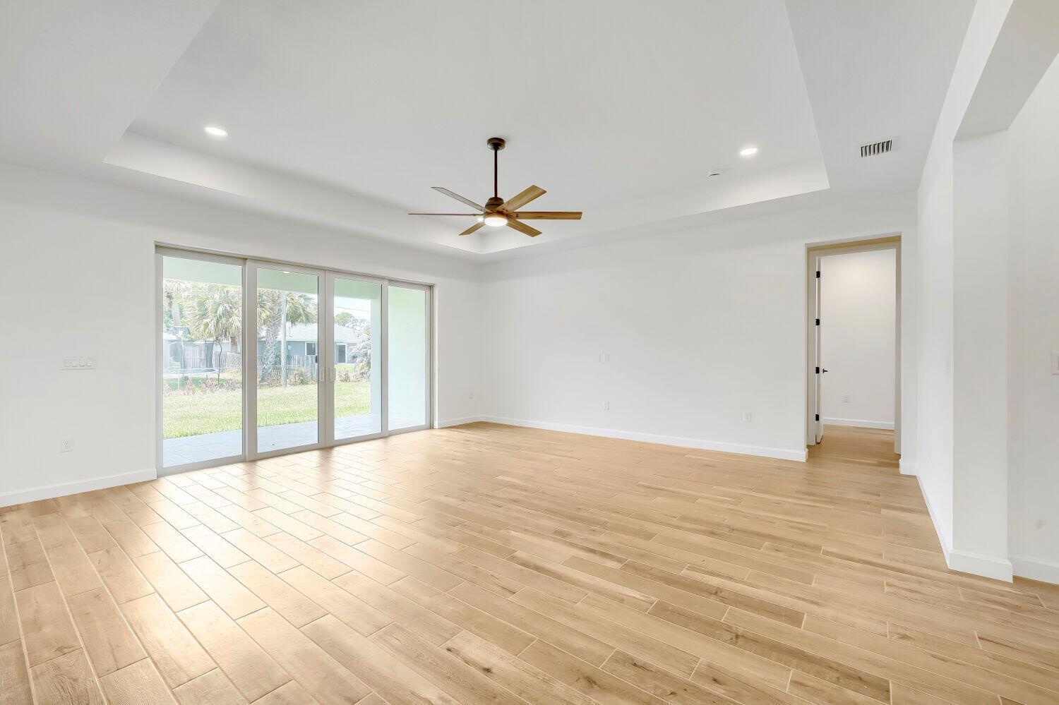 A bright, empty living room features light wood-look tile flooring, a tray ceiling, and sliding doors — 6635 49th Court, Vero Beach FL 32967, VERO BEACH, FL