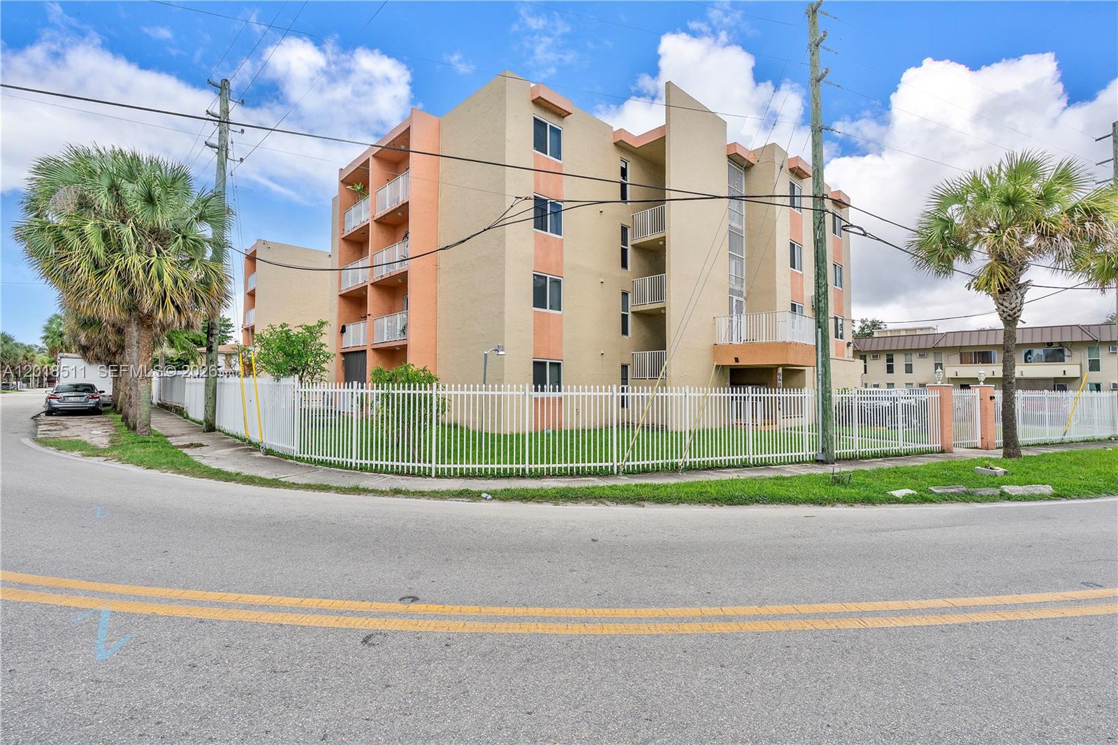 A multi-story apartment building with balconies stands behind a white fence near a road — 2400 NW 16th St Rd, Miami FL 33125, MIAMI, FL