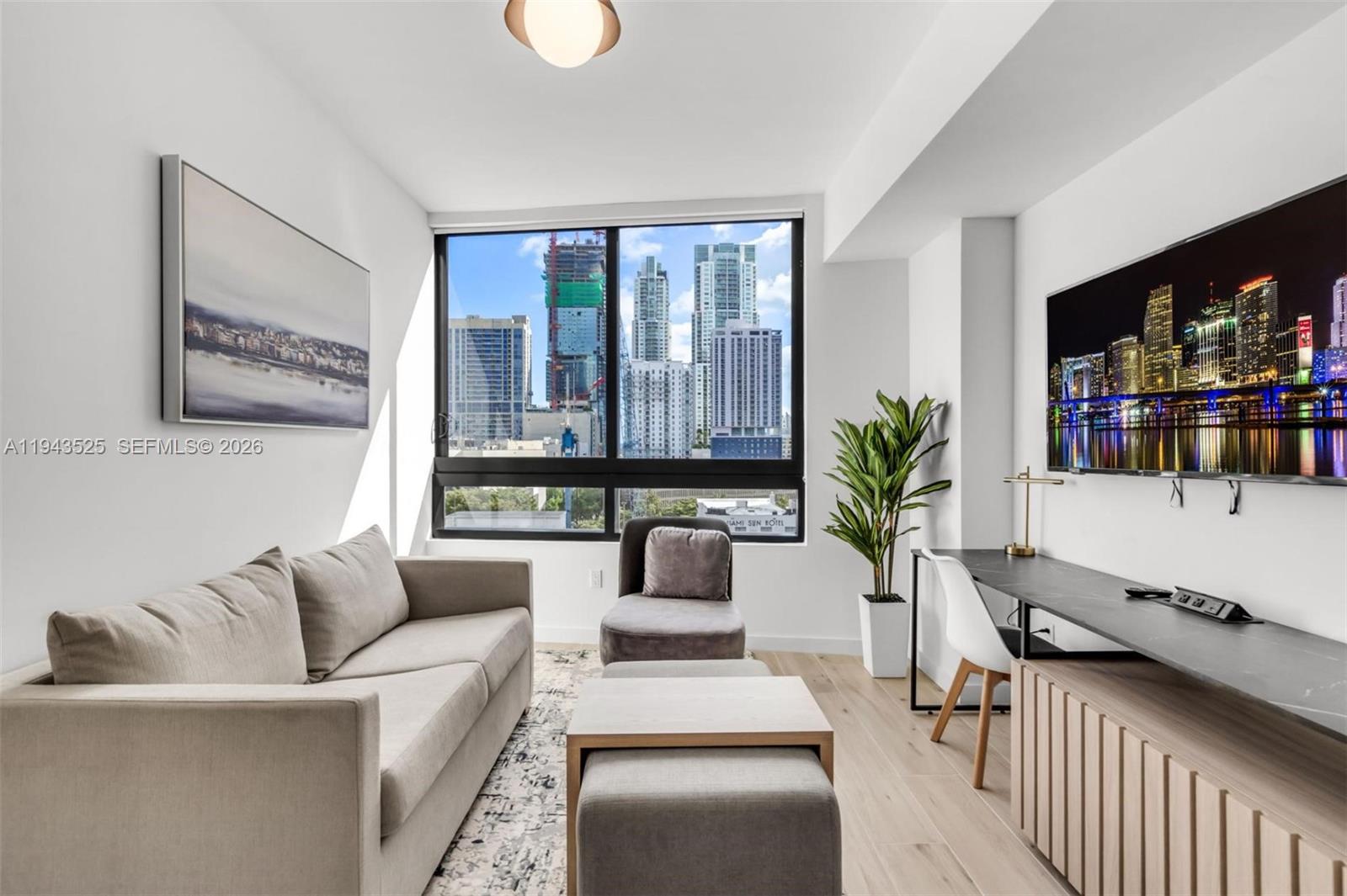 A modern living room features a sofa, desk, television, and large windows overlooking city skyscrapers — 225 N Miami Ave # 1005, Miami FL 33128, MIAMI, FL