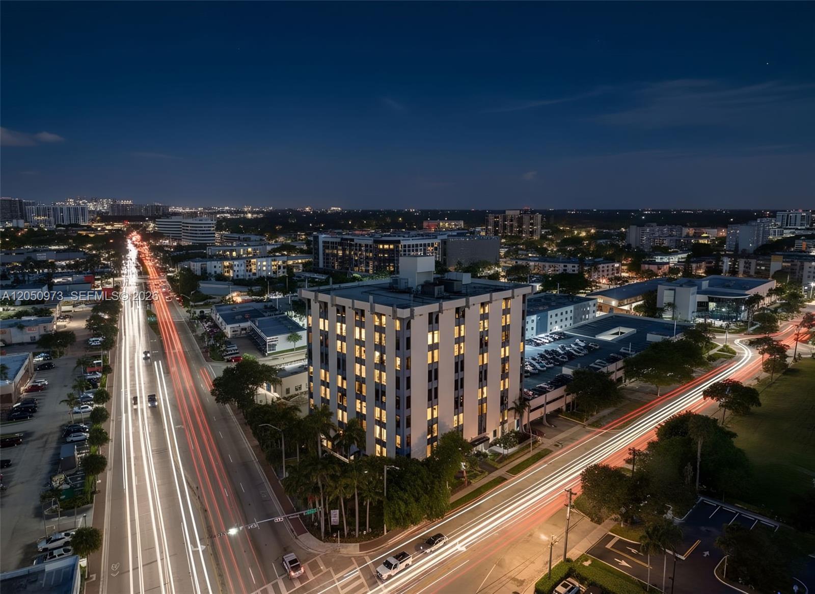 An aerial night view shows a multi-story building surrounded by illuminated streets with light trails — 12550 Biscayne Blvd # 702, North Miami FL 33181, NORTH MIAMI, FL