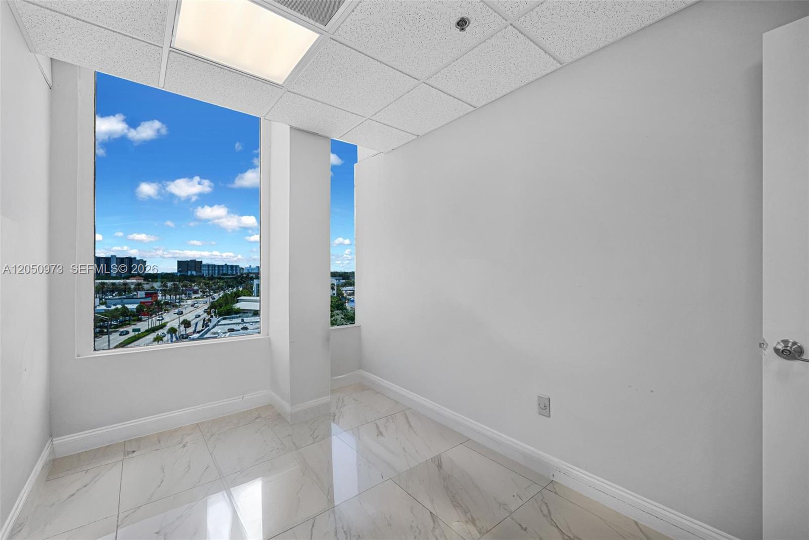 An empty office with white walls, marble-patterned tile floors, and a large window overlooking city streets — 12550 Biscayne Blvd # 702, North Miami FL 33181, NORTH MIAMI, FL