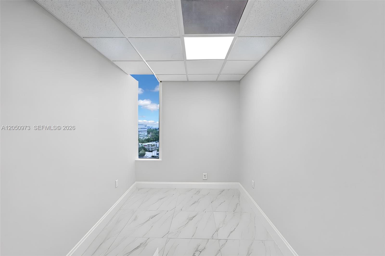 An empty office space features white walls, marble-patterned tile flooring, and a tall window — 12550 Biscayne Blvd # 702, North Miami FL 33181, NORTH MIAMI, FL