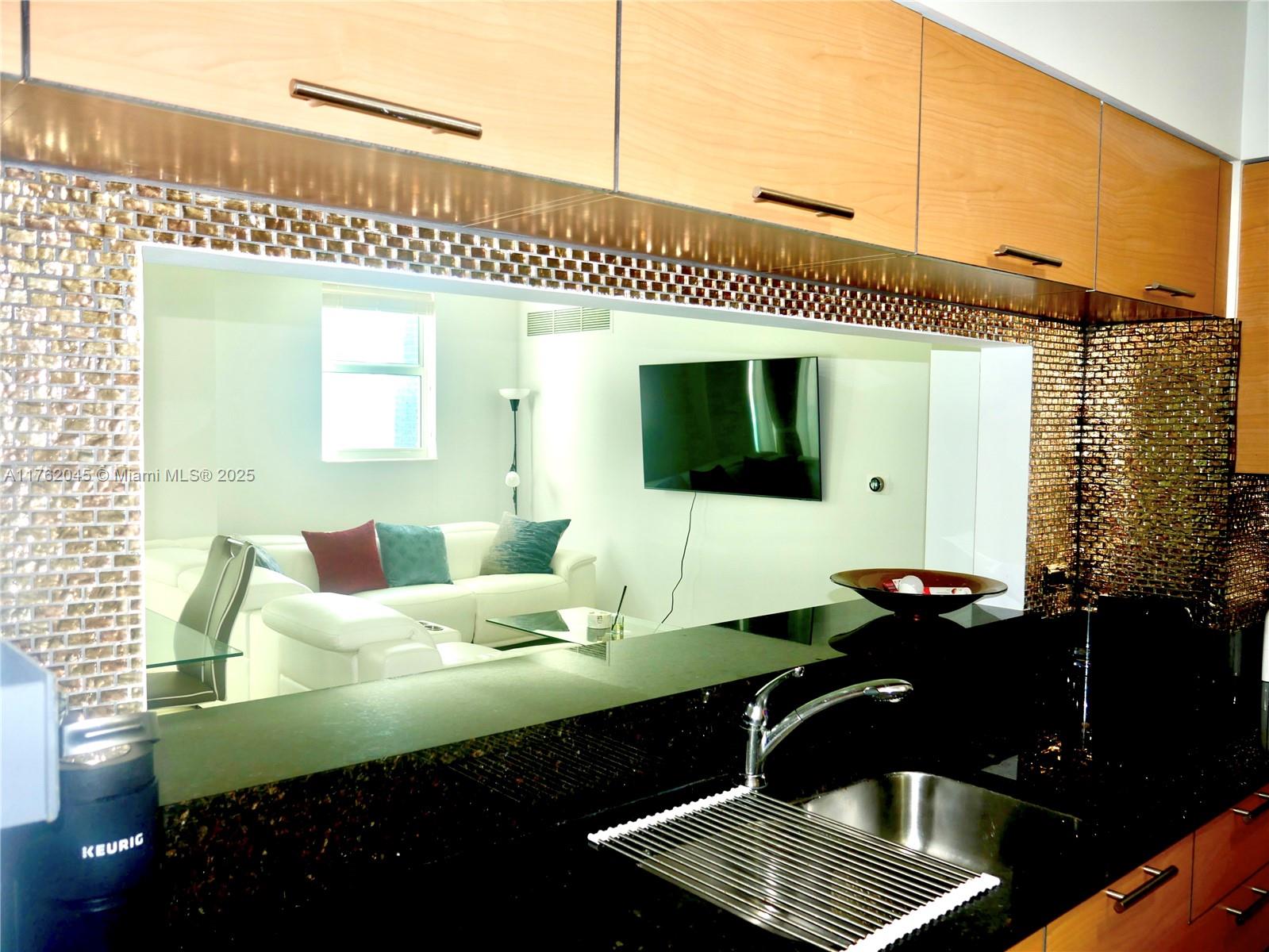 A kitchen with dark countertops and wood cabinets overlooks a living room through a window — 31 SE 5th St # 2611, Miami FL 33131, MIAMI, FL