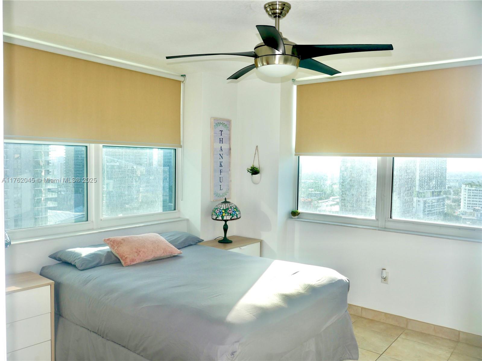 A bedroom features a bed, a ceiling fan, and large windows with tan roller shades — 31 SE 5th St # 2611, Miami FL 33131, MIAMI, FL