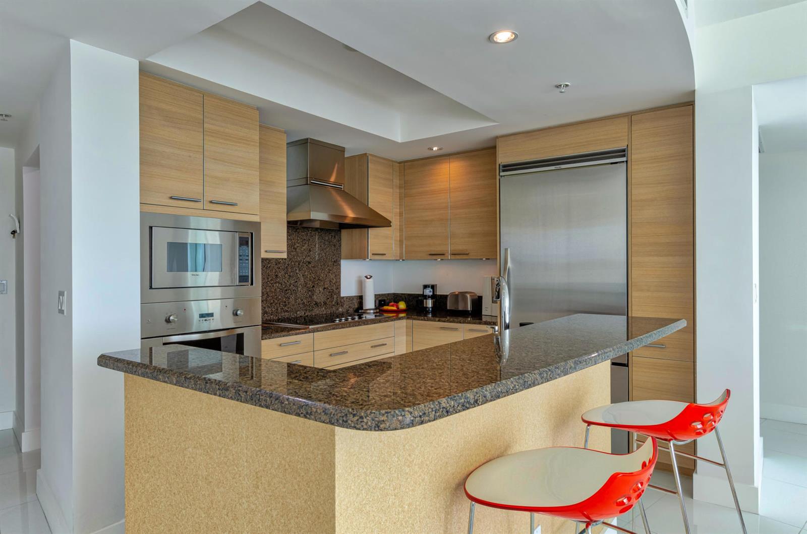 A modern kitchen features light wood cabinetry, granite countertops, stainless steel appliances, and two red stools — 1 N Ocean Boulevard # 913, Pompano Beach FL 33062, POMPANO BEACH, FL