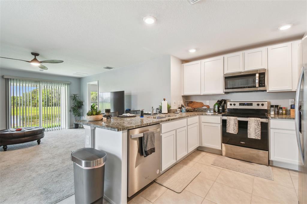 A kitchen with white cabinets, granite countertops, and stainless steel appliances overlooks a carpeted living area — 36673 SPANISH ROSE DRIVE, DADE CITY, FL