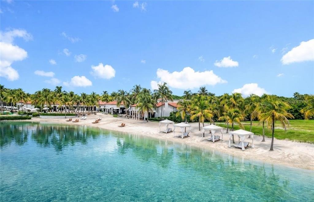 A sandy beach with cabanas and palm trees sits in front of a resort building — 10264 PORTO ROMANO DRIVE, MIROMAR LAKES, FL