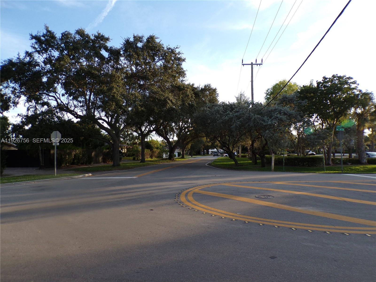 An asphalt intersection features yellow painted road markings and mature trees under a blue sky — 5030 N Federal Hwy, Lighthouse Point FL 33064, LIGHTHOUSE POINT, FL