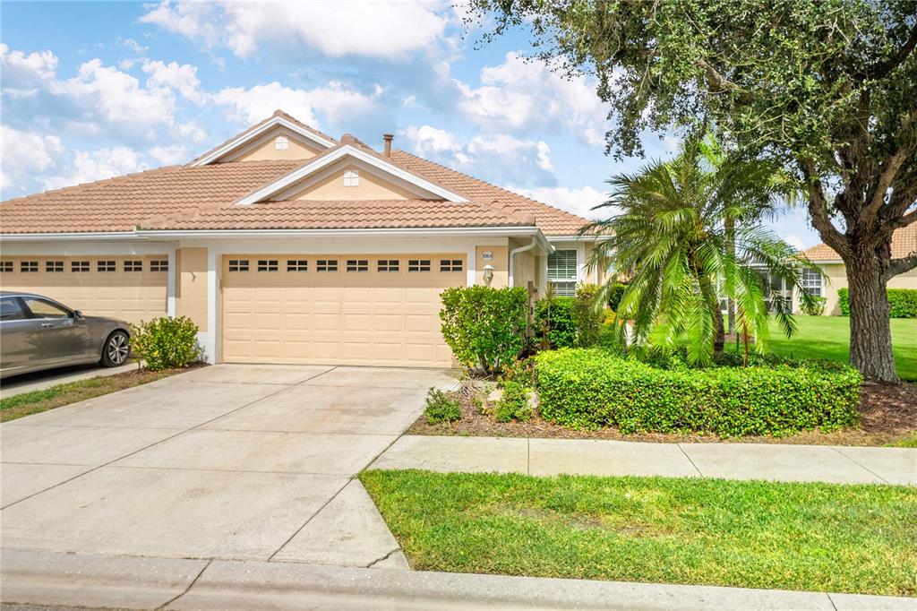 A beige single-story house features a two-car garage, a tiled roof, and landscaped front yard — 5064 WHISPERING OAKS DRIVE, NORTH PORT, FL