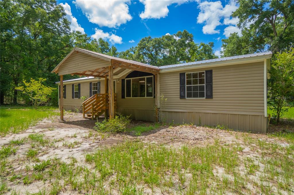 A tan manufactured home with a covered front porch sits on a grassy, wooded lot — 2349 NE 46TH CIRCLE, HIGH SPRINGS, FL