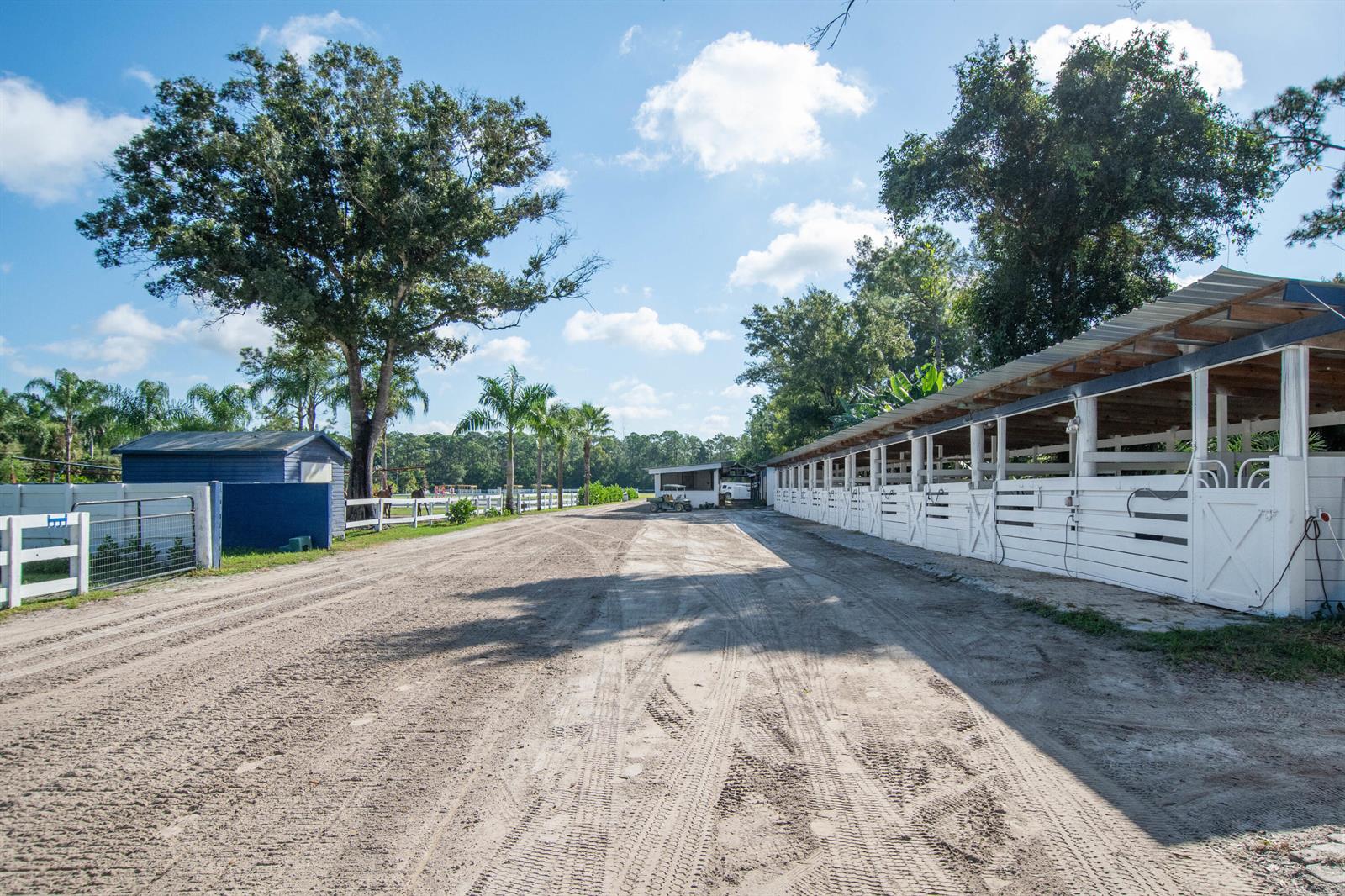 A dirt path leads between a long white horse stable and a blue storage shed — 1044 D Road, FL 33470, FL