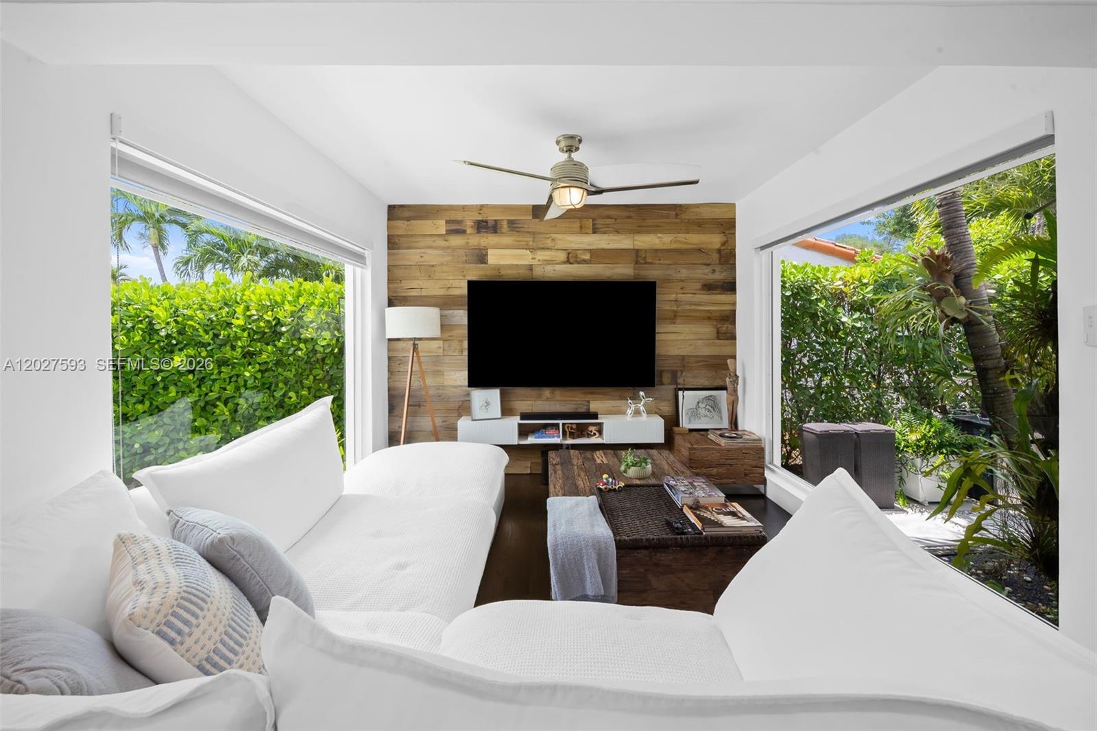A living room features a wood-paneled accent wall, a television, and large windows overlooking greenery — 39 Montilla Ave, Coral Gables FL 33134, CORAL GABLES, FL