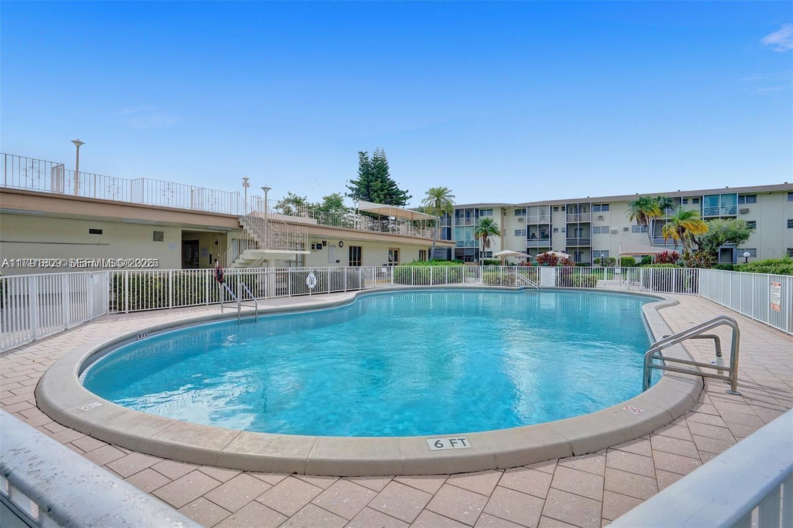 An outdoor swimming pool is surrounded by a white fence and a multi-story apartment building — 1355 NE 167th St # 302, Miami FL 33162, MIAMI, FL