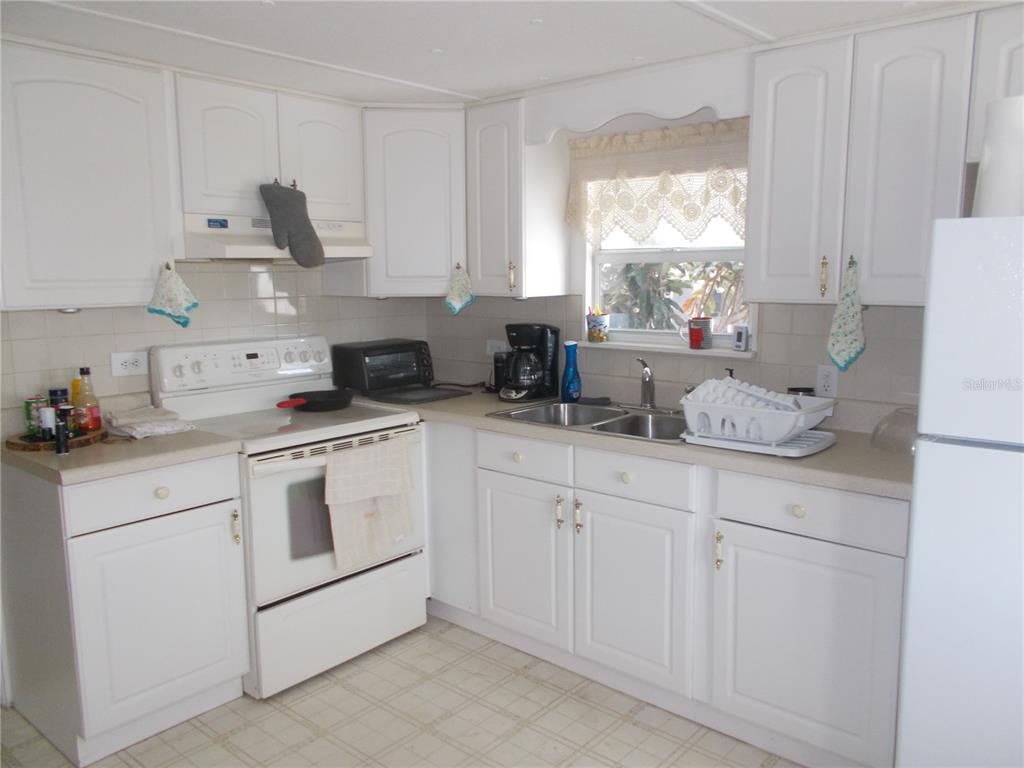 A kitchen with white cabinets, white appliances, a double sink, and a window with curtains — 6427 FLEETWOOD COURT, NORTH PORT, FL