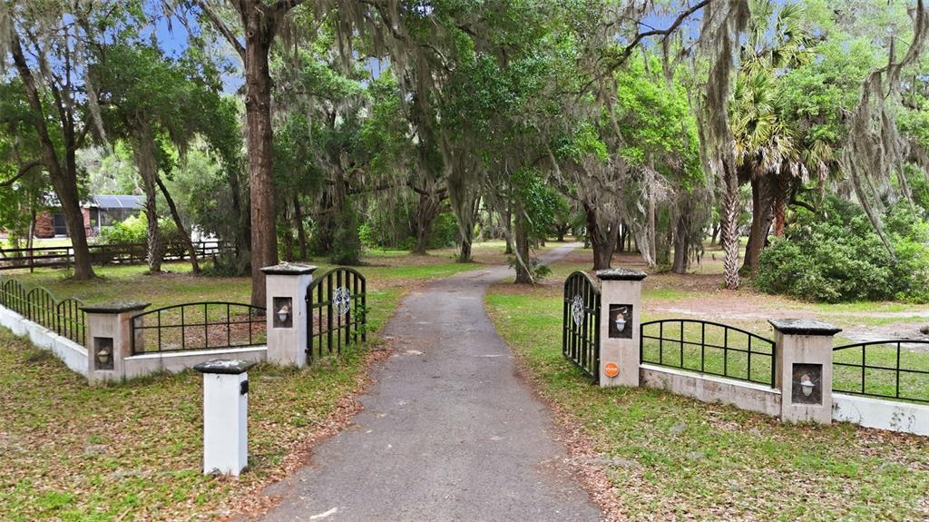 A paved driveway leads through an open metal gate flanked by stone pillars and fencing — 10639 SE SUNSET HARBOR ROAD, SUMMERFIELD, FL