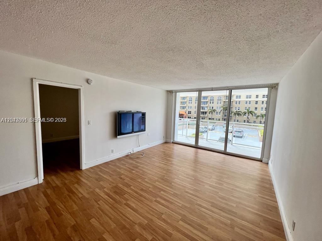 An empty living room features wood-look flooring, a wall-mounted television, and a large sliding glass door — 2030 S Ocean Dr # 211, Hallandale Beach FL 33009, HALLANDALE BEACH, FL