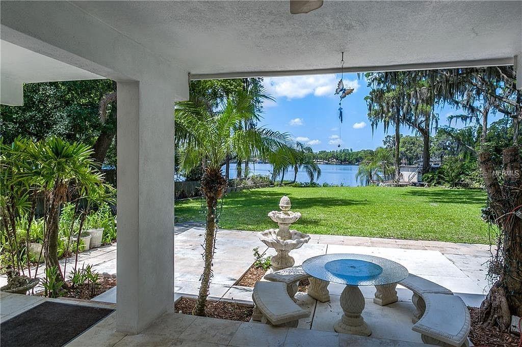 A covered patio overlooks a grassy lawn, a fountain, and a lake with trees — 18618 WAYNE ROAD, ODESSA, FL