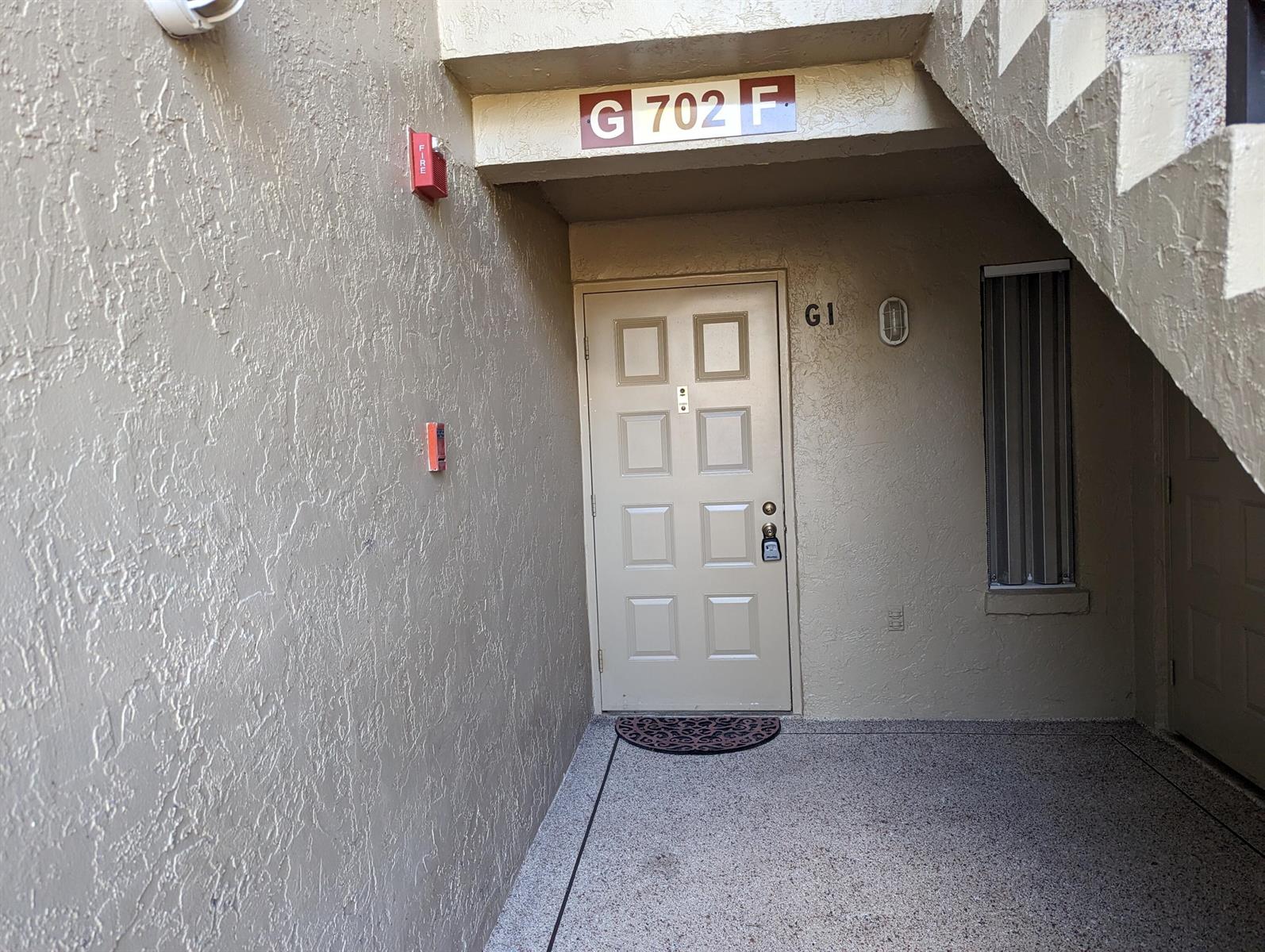 A beige apartment entrance door with a lockbox sits under a stairwell overhang — 702 Sunny Pine Way # G1, FL 33415, FL