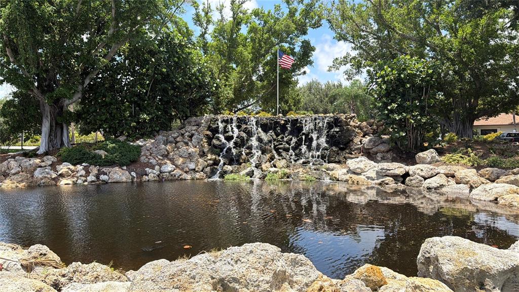 A stone waterfall feature flows into a pond surrounded by trees and an American flag — 622 BIRD BAY DRIVE S UNIT 203, VENICE, FL