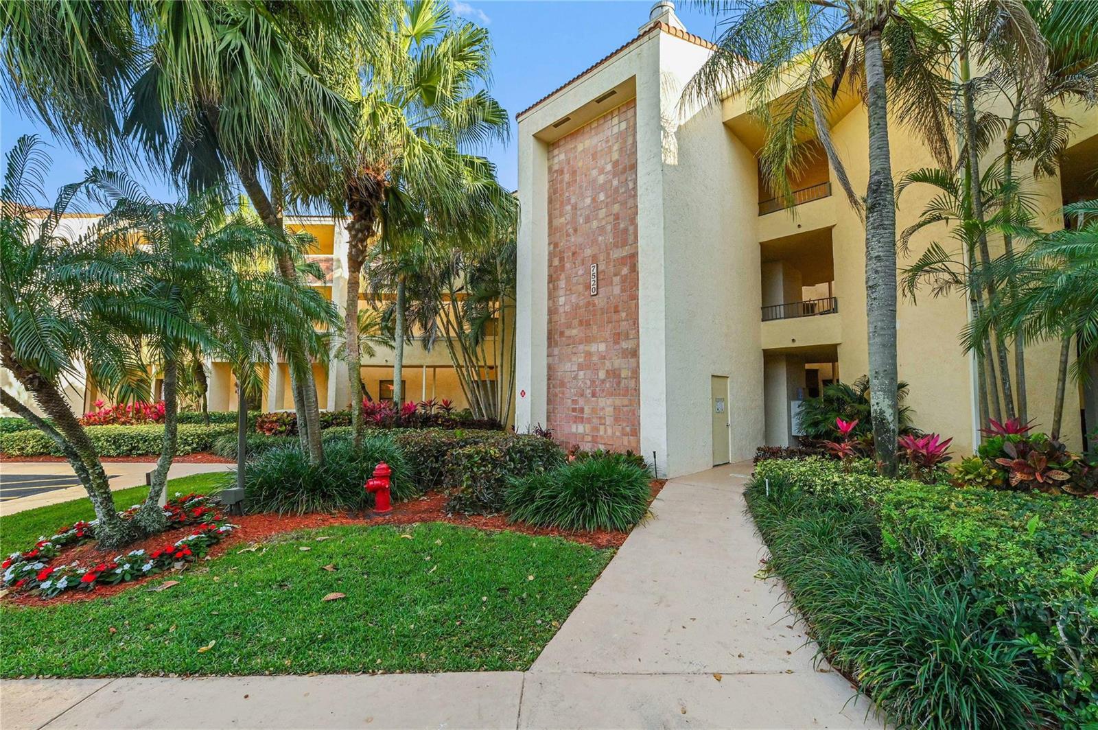 A concrete walkway leads to the entrance of a multi-story apartment building with palm trees — 7520 La Paz Boulevard # 306, Boca Raton FL 33433, BOCA RATON, FL
