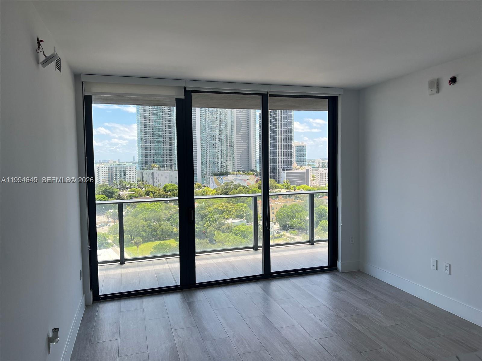 An empty room with light wood-look flooring features a balcony with a city skyline view — 121 NE 34th St # 1207, Miami FL 33137, MIAMI, FL
