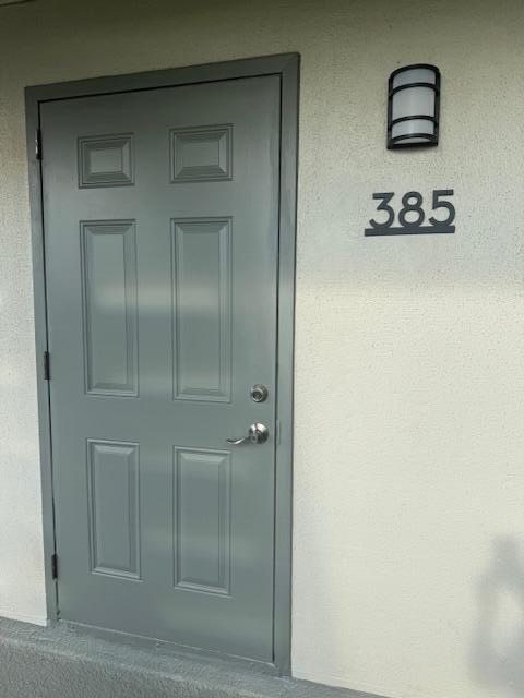 A gray six-panel door with a silver handle sits beside a wall-mounted light and address numbers — 385 Flanders I, Delray Beach FL 33484, DELRAY BEACH, FL