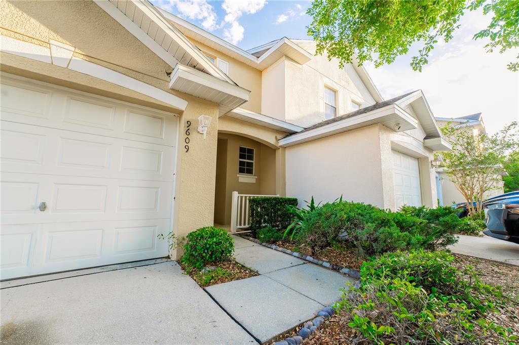 A two-story house exterior features a white garage door, a covered entryway, and landscaping — 9609 CARLSDALE DRIVE, RIVERVIEW, FL