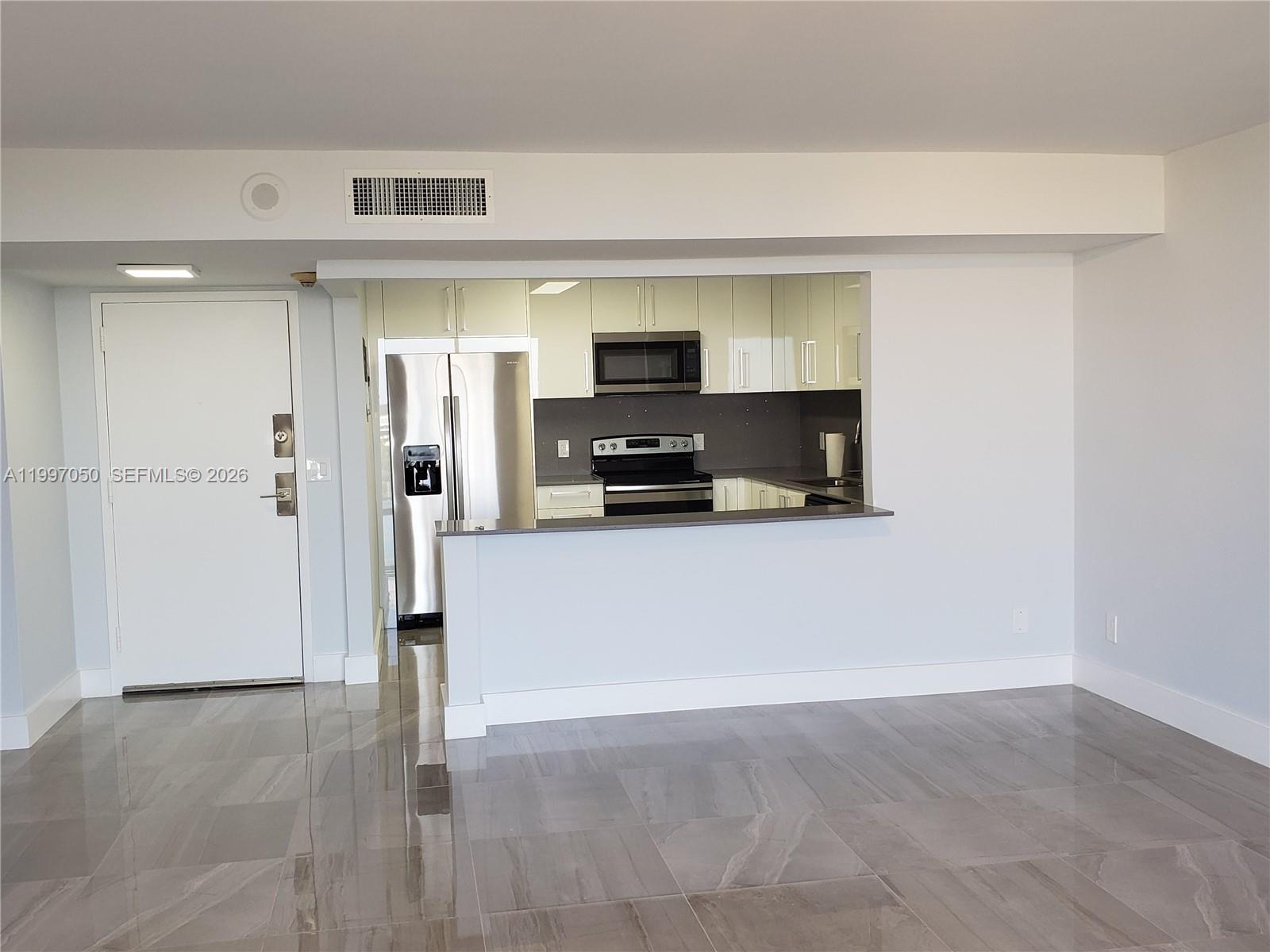 A kitchen with stainless steel appliances and white cabinetry overlooks a room with tile flooring — 13499 Biscayne Blvd # 1105, North Miami FL 33181, NORTH MIAMI, FL