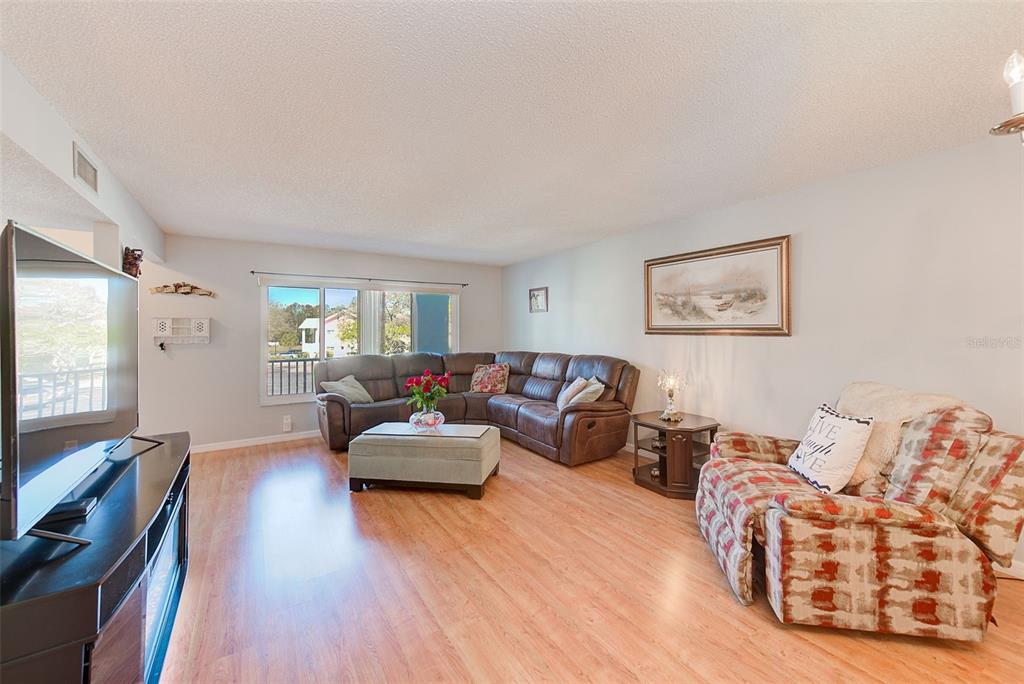 A living room features a brown sectional sofa, patterned armchair, and light wood-look flooring — 2229 AMERICUS BOULEVARD W UNIT 25, CLEARWATER, FL
