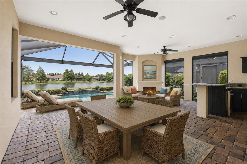 A covered patio features a dining set, fireplace, outdoor kitchen, and a pool overlooking a lake — 12749 FONTANA LOOP, BRADENTON, FL