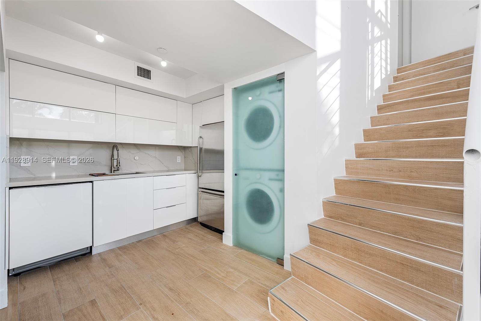 A modern kitchen with white cabinetry and a stacked laundry unit next to wooden stairs — 221 Jefferson Ave # 2, Miami Beach FL 33139, MIAMI BEACH, FL