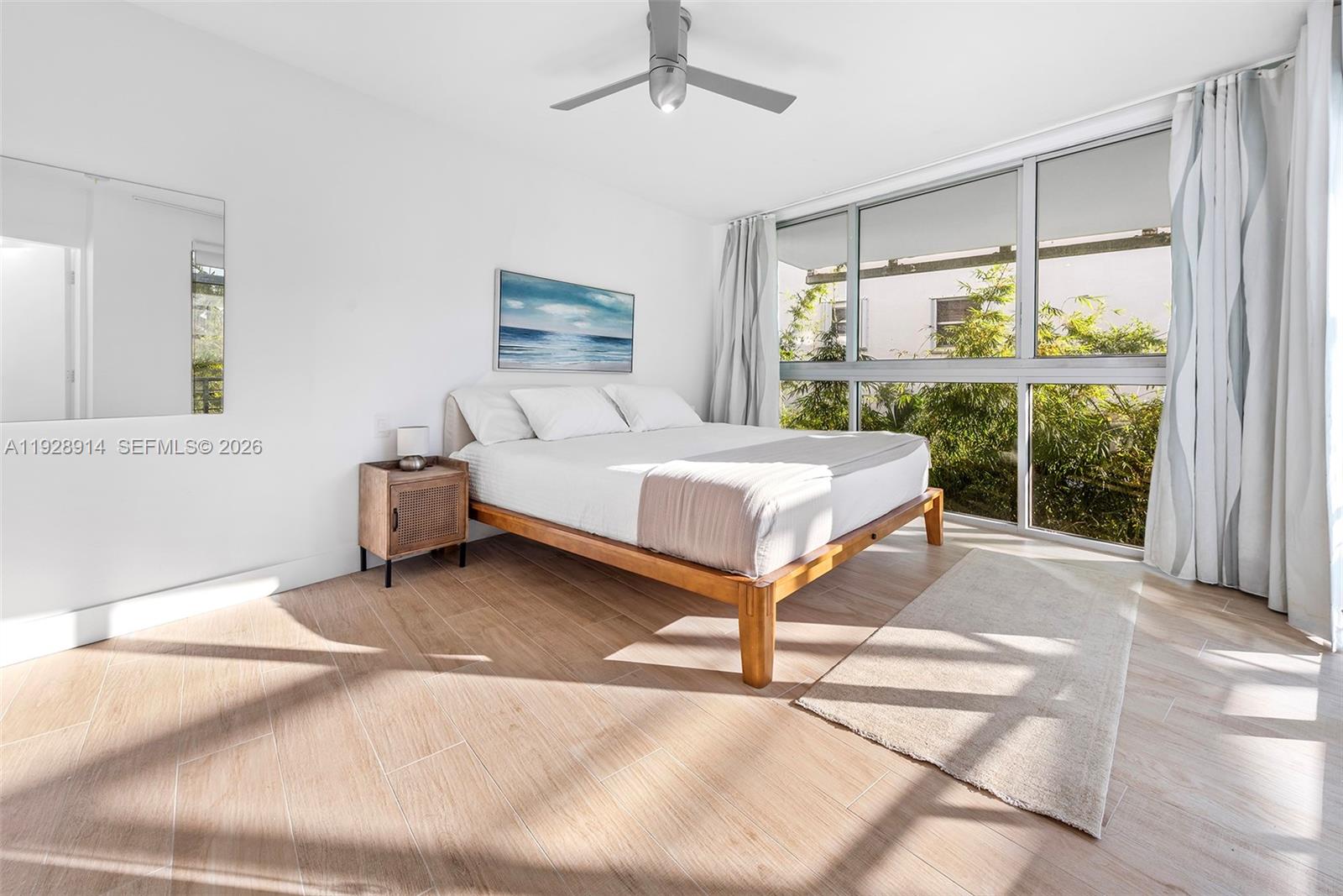 A bedroom features a wooden bed frame, light wood-look tile flooring, and large floor-to-ceiling windows — 221 Jefferson Ave # 2, Miami Beach FL 33139, MIAMI BEACH, FL
