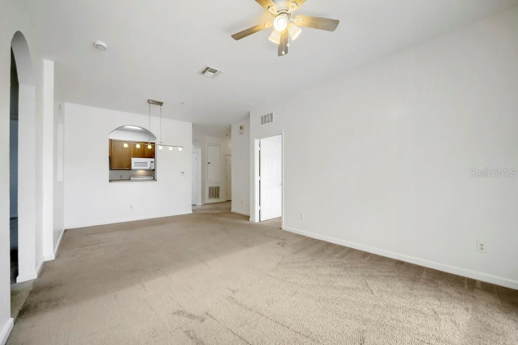 A spacious living room with beige carpeting, a ceiling fan, and an arched kitchen pass-through — 6466 CAVA ALTA DRIVE UNIT 208, ORLANDO, FL