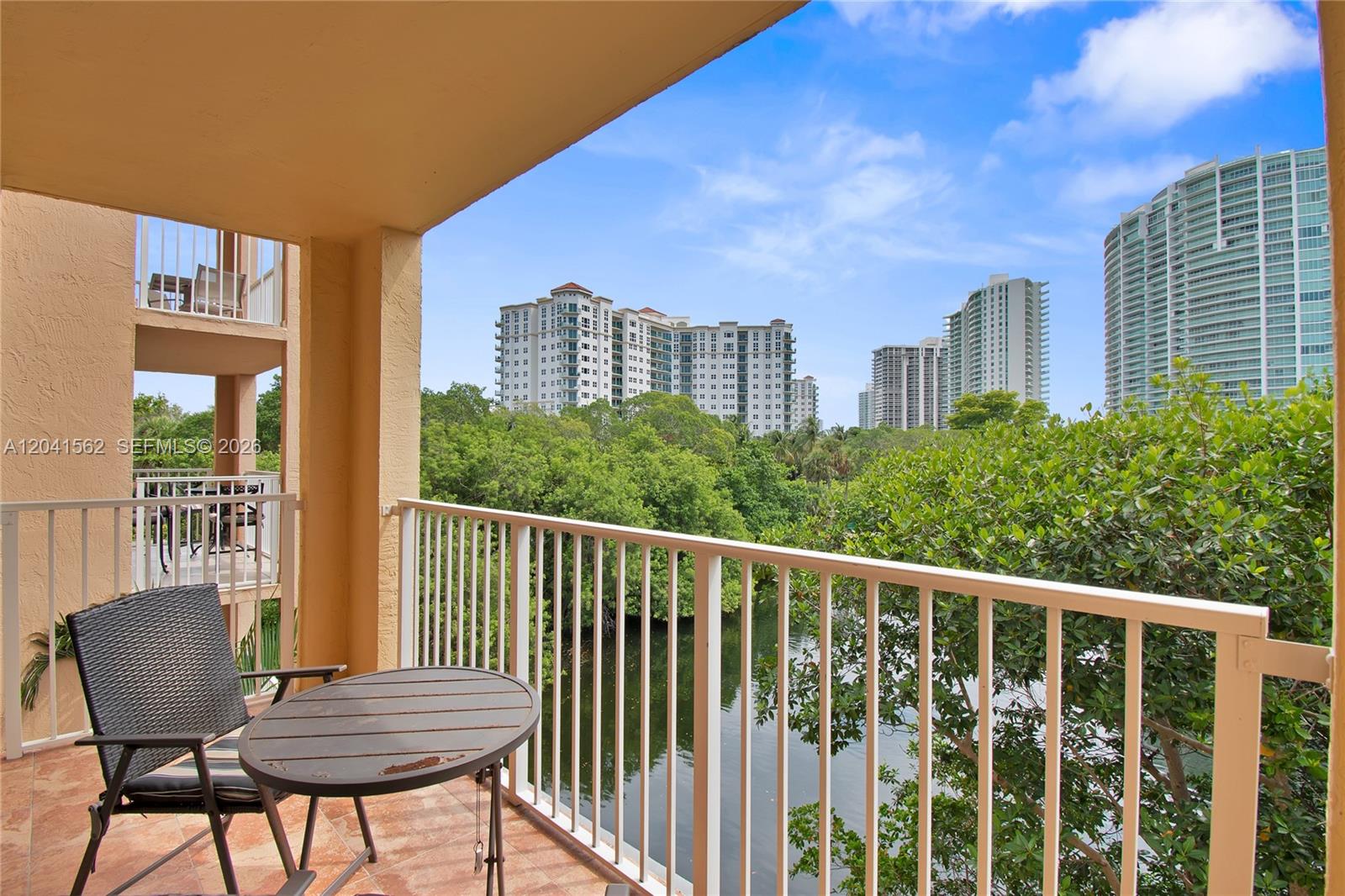 A balcony with a table and chair overlooks a canal, trees, and high-rise buildings — AVENTURA, FL