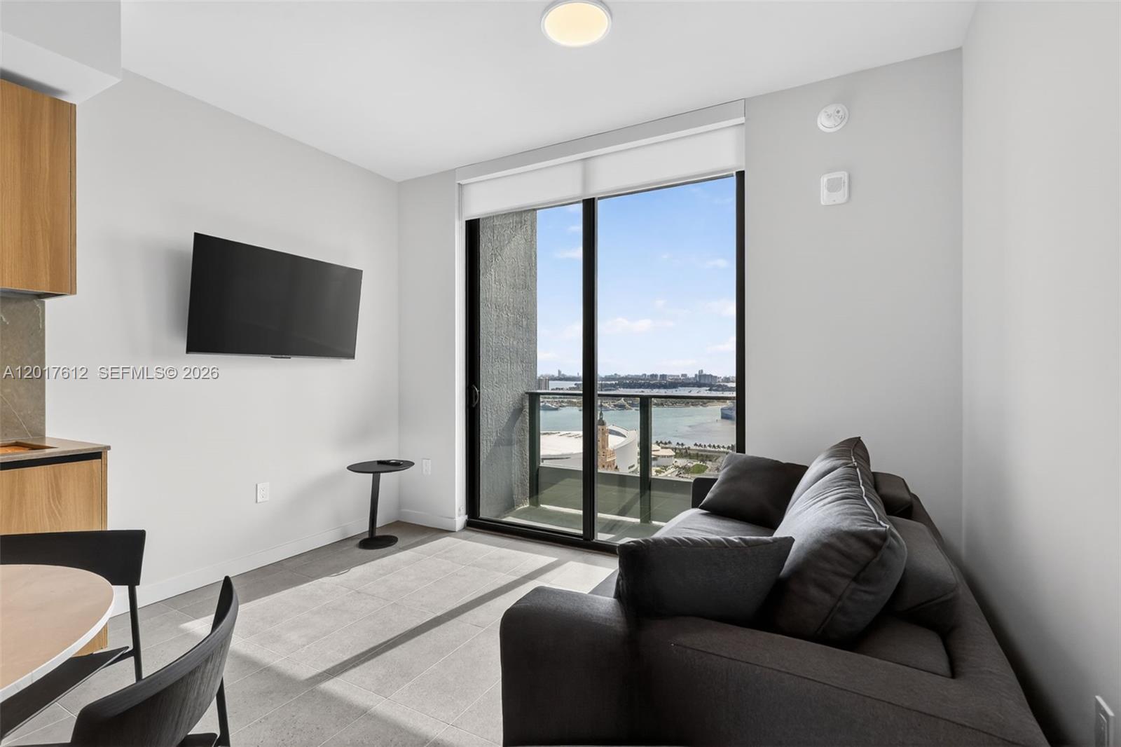 A modern living room features a dark sofa, wall-mounted television, and a balcony with water views — 121 NE 5th St # 3313, Miami FL 33132, MIAMI, FL