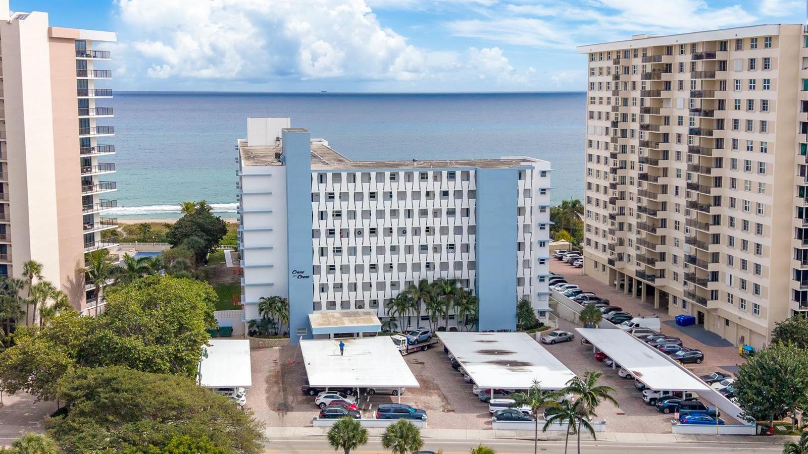 An aerial view shows a blue beachfront apartment building with covered parking lots below — 1850 S Ocean Boulevard # 502, Pompano Beach FL 33062, POMPANO BEACH, FL