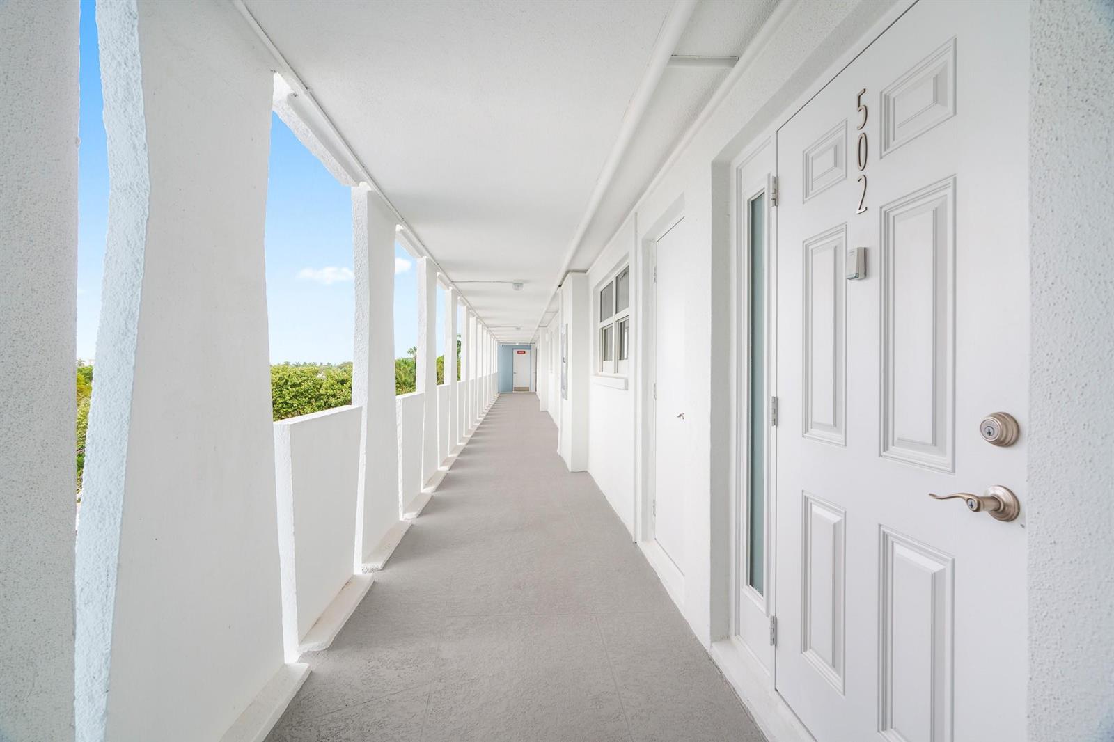 An outdoor apartment hallway with white walls, a gray floor, and a view of trees — 1850 S Ocean Boulevard # 502, Pompano Beach FL 33062, POMPANO BEACH, FL