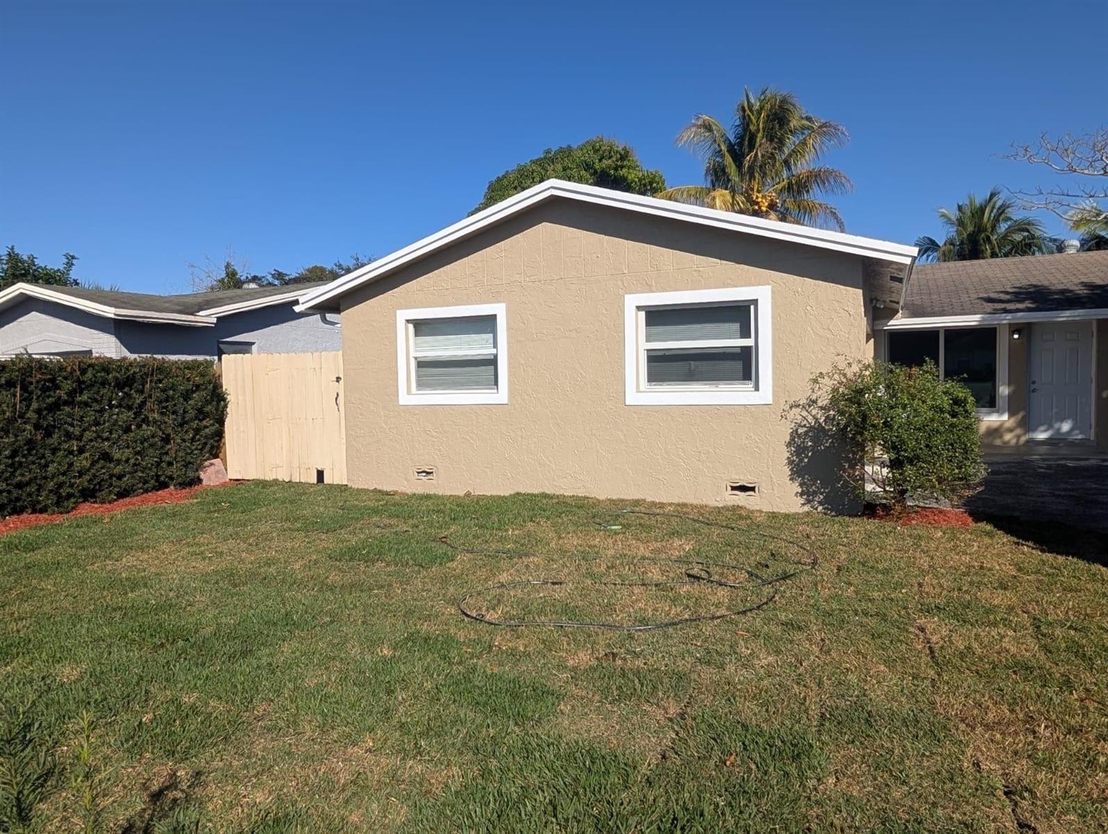 The exterior of a tan, single-story house features two windows and a wooden fence — 7713 Kimberly Boulevard, North Lauderdale FL 33068, NORTH LAUDERDALE, FL