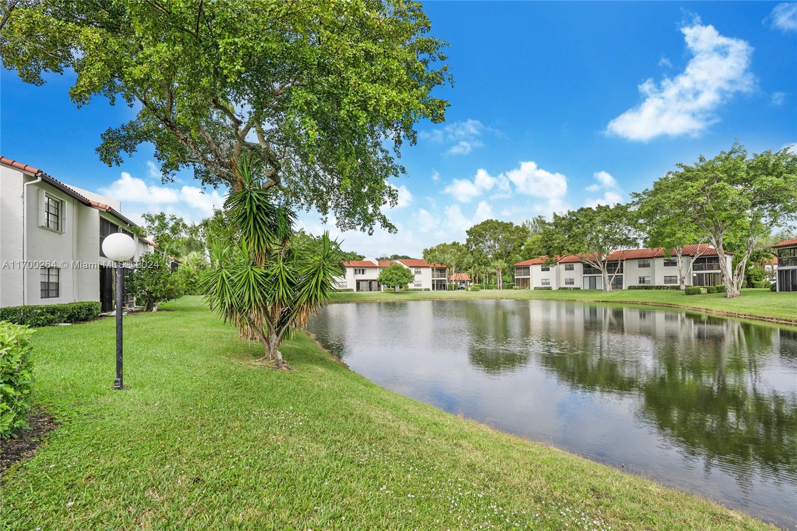 A pond sits in front of multi-story apartment buildings surrounded by green lawns and trees — 21364 Cypress Hammock Dr # 1H, Boca Raton FL 33428, BOCA RATON, FL