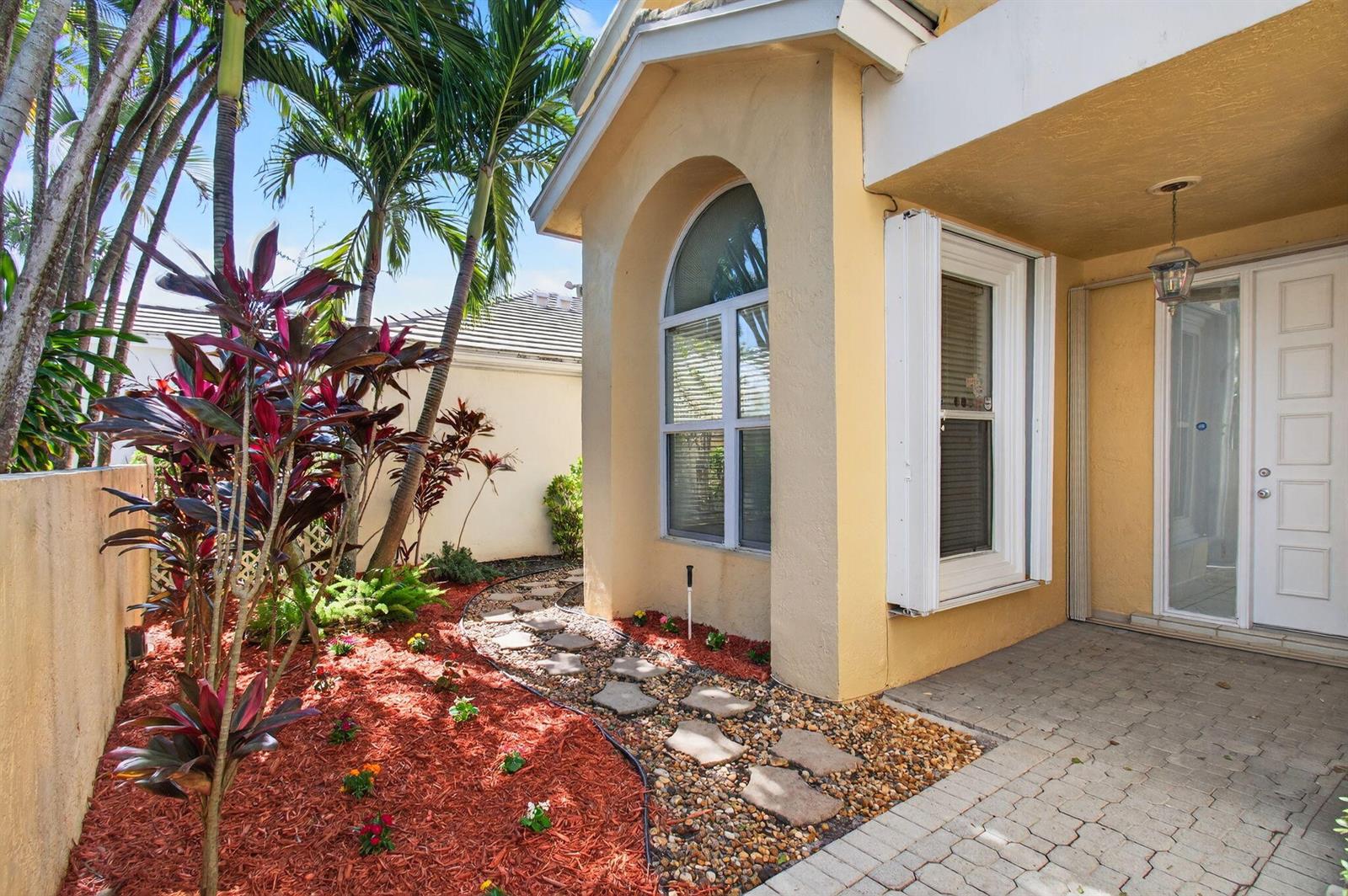 A paved walkway leads to the front entrance of a yellow house with tropical landscaping — 7848 Travlers Tree Drive, Boca Raton FL 33433, BOCA RATON, FL