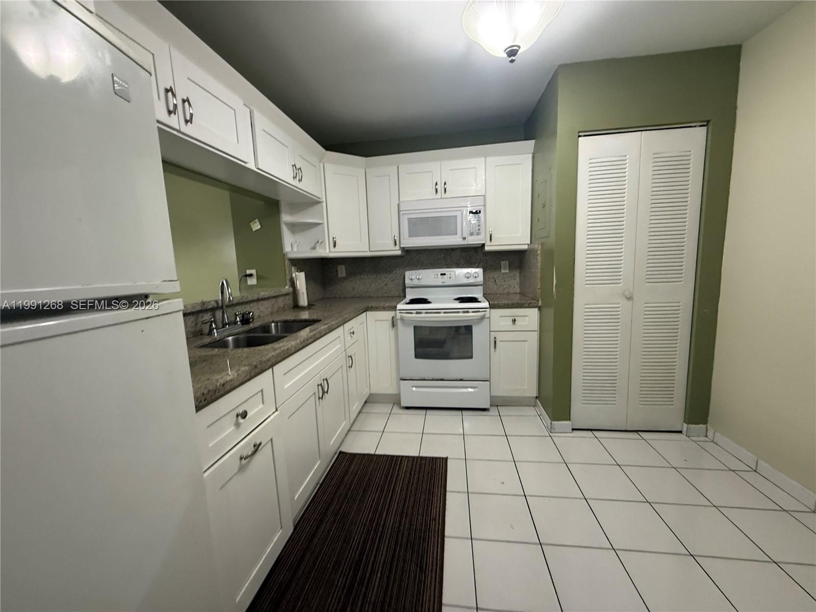 A kitchen features white cabinets, granite countertops, white appliances, and white tile flooring — 1800 W 54th St # 108, Hialeah FL 33012, HIALEAH, FL