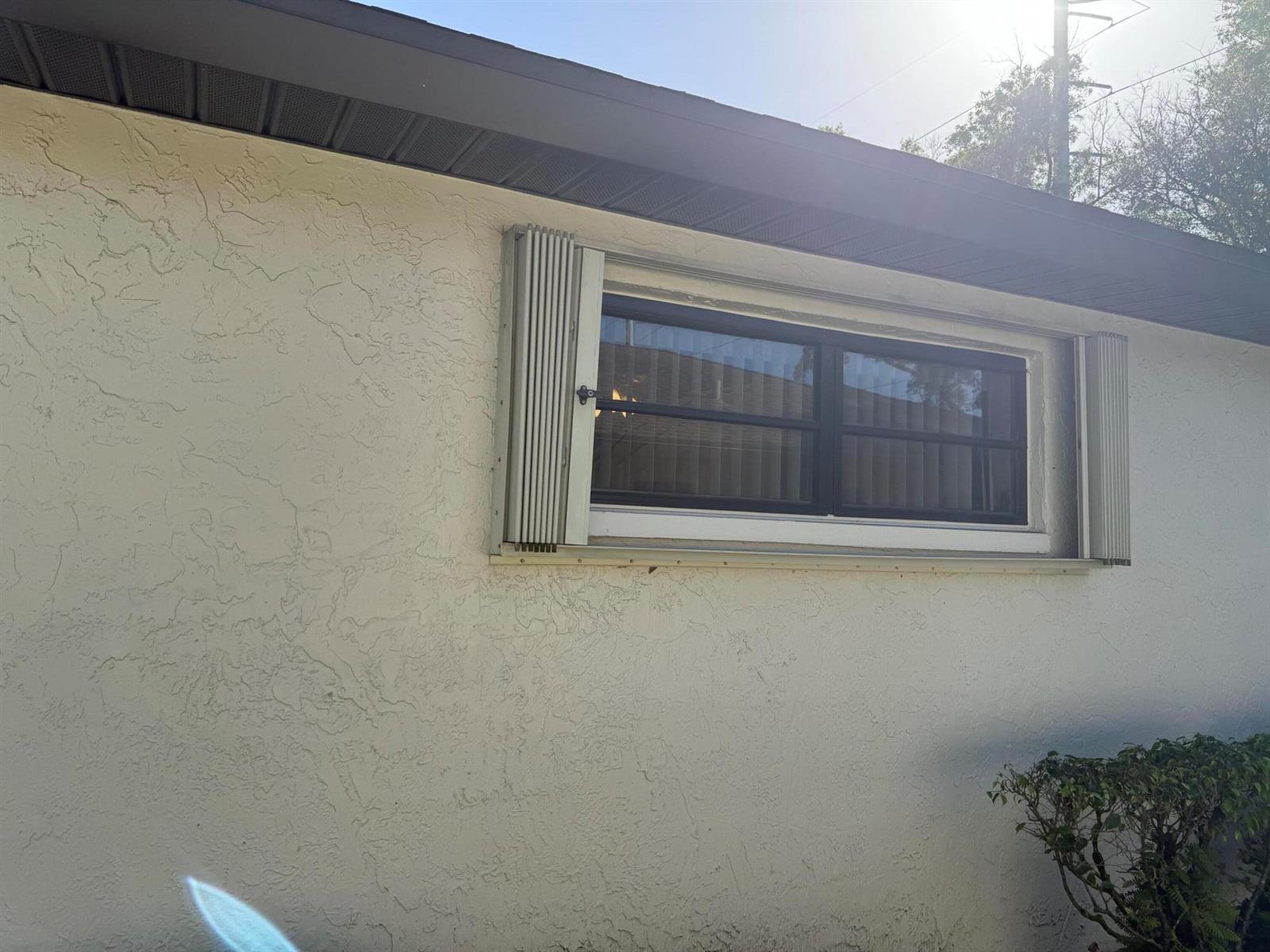 An exterior view of a stucco house wall featuring a window with accordion hurricane shutters — 4948 Equestrian Circle # B, Boynton Beach FL 33436, BOYNTON BEACH, FL