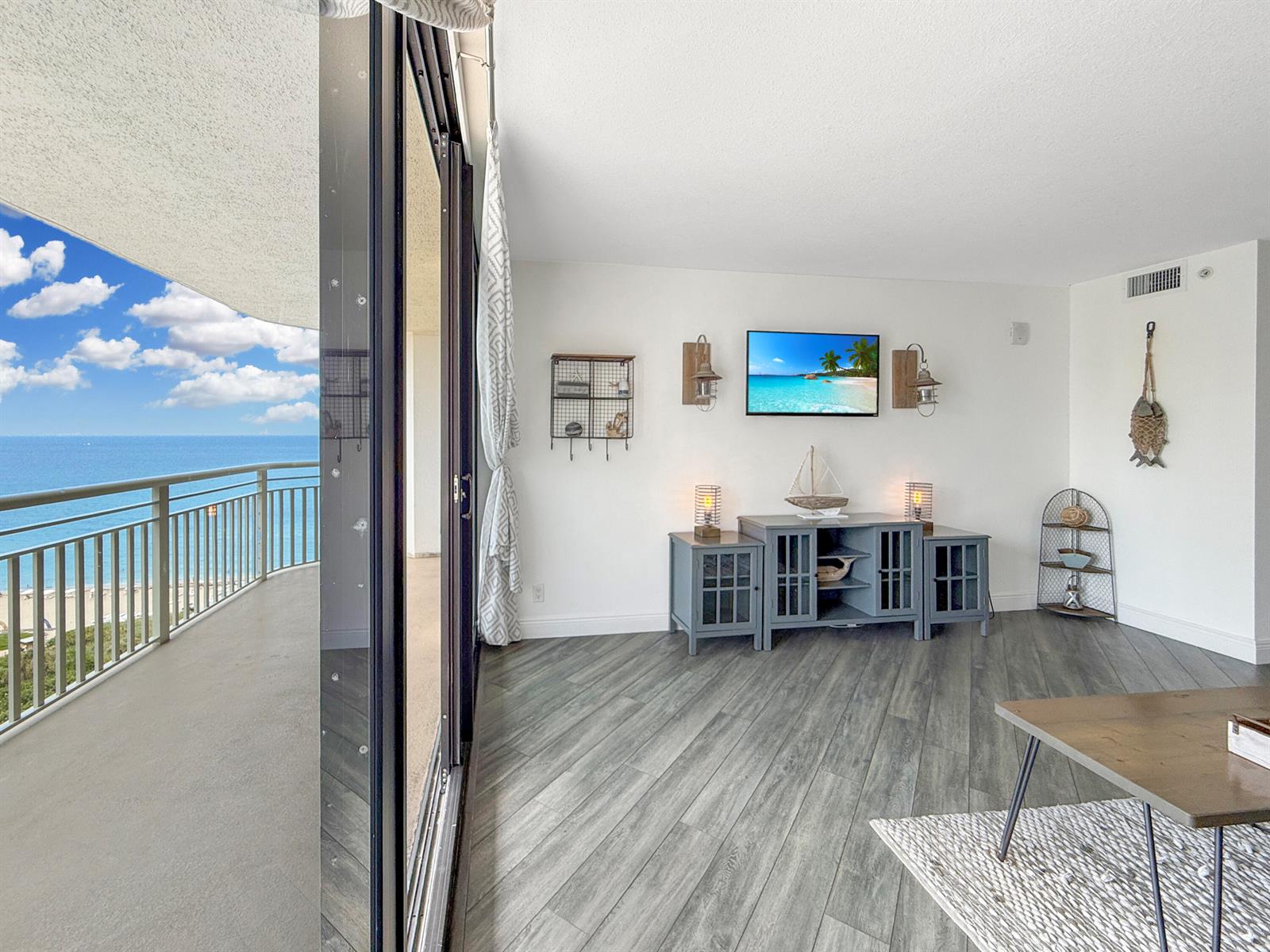 A living room features gray wood-look flooring, a wall-mounted television, and a balcony overlooking the ocean — 3000 N Ocean Drive # B, Singer Island FL 33404, SINGER ISLAND, FL